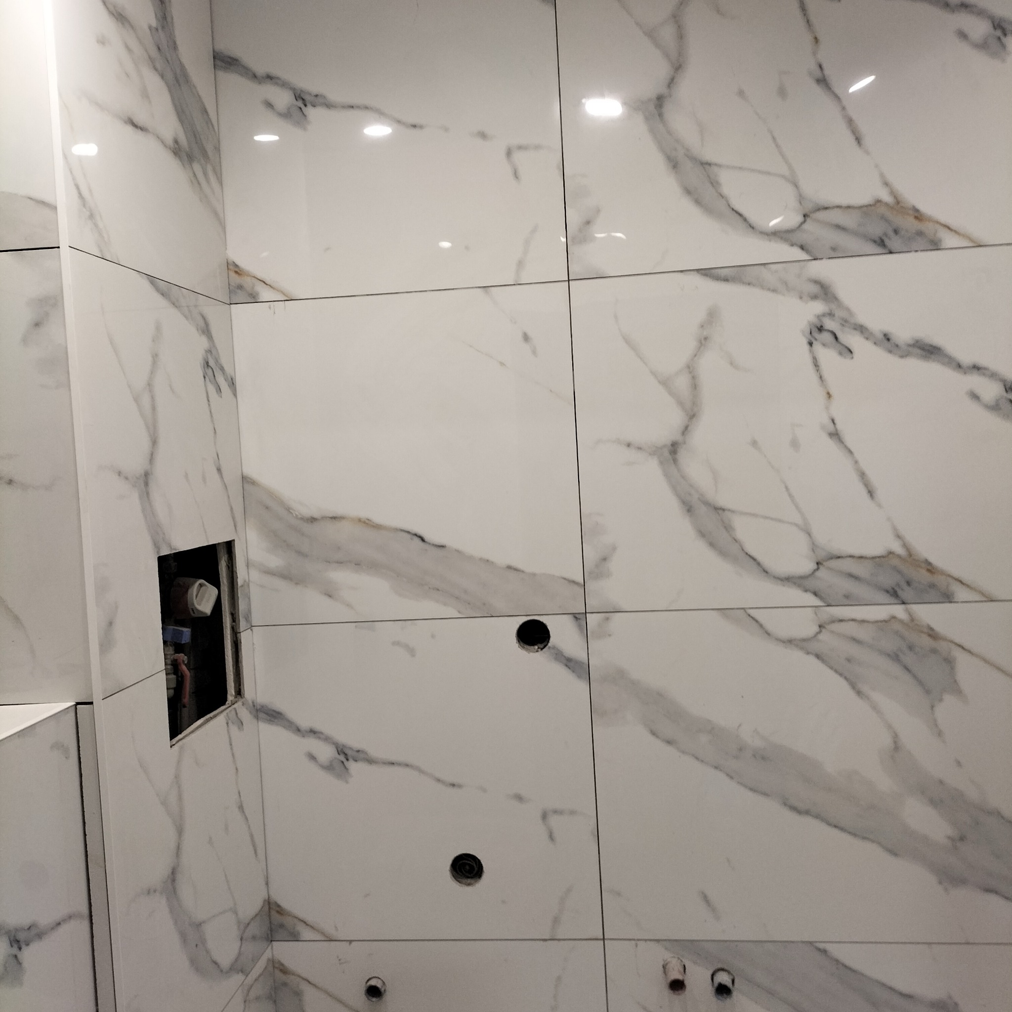 Ściana łazienki wyłożona białymi płytkami imitującymi marmur, z widocznymi otworami na instalacje i puszkę instalacyjną w trakcie montażu.