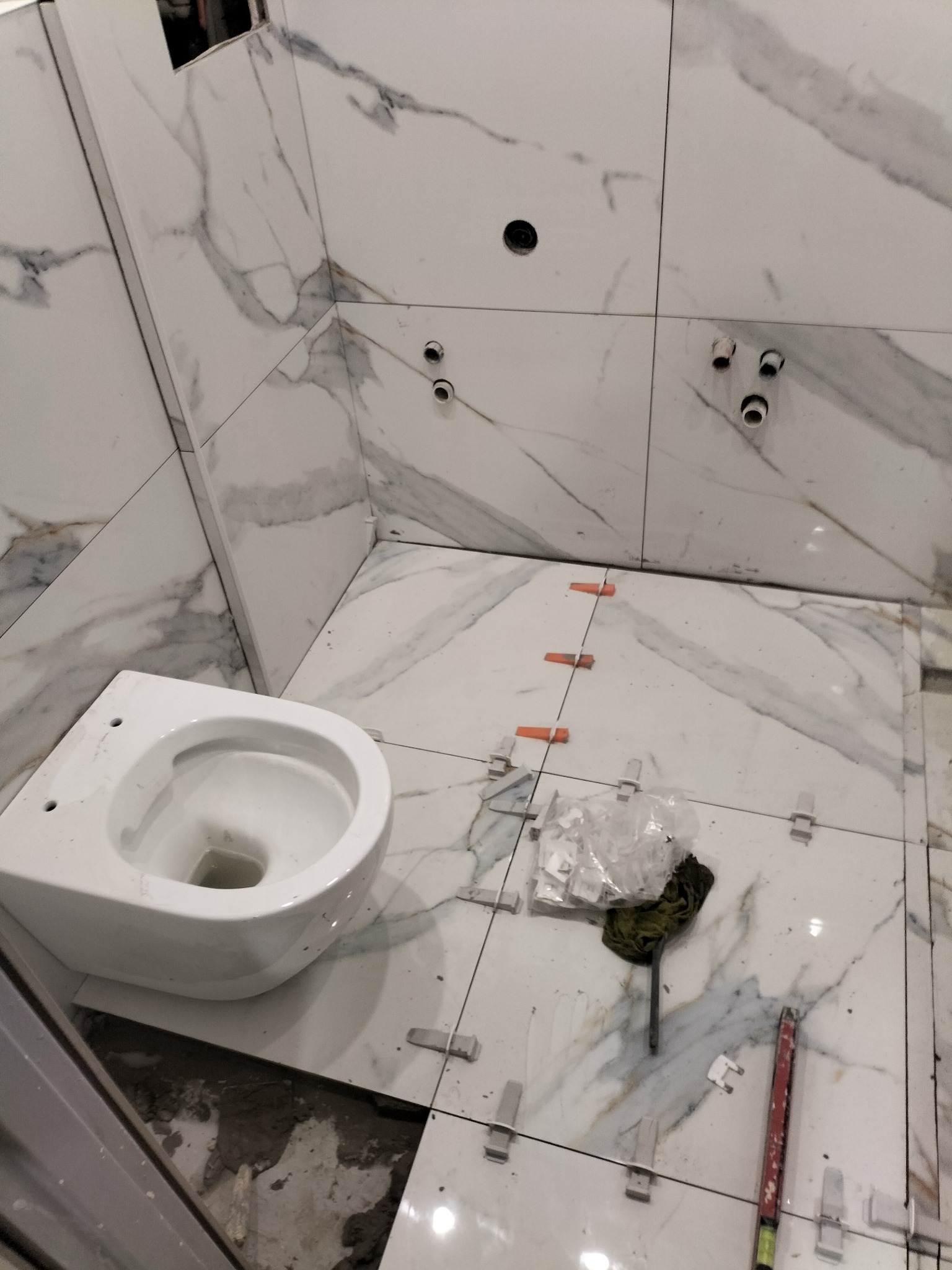 Biała, wisząca muszla klozetowa w trakcie montażu w łazience z marmurowymi płytkami. Widoczne akcesoria glazurnicze, takie jak krzyżyki dystansowe i poziomica.