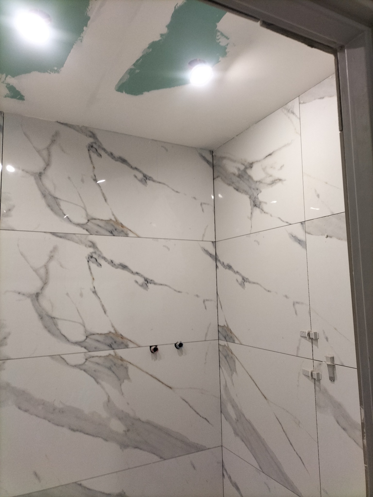 Fragment łazienki w trakcie prac: białe płytki imitujące marmur na ścianach, widoczne krzyżyki glazurnicze, sufit w trakcie szpachlowania, instalacja wodna przygotowana.