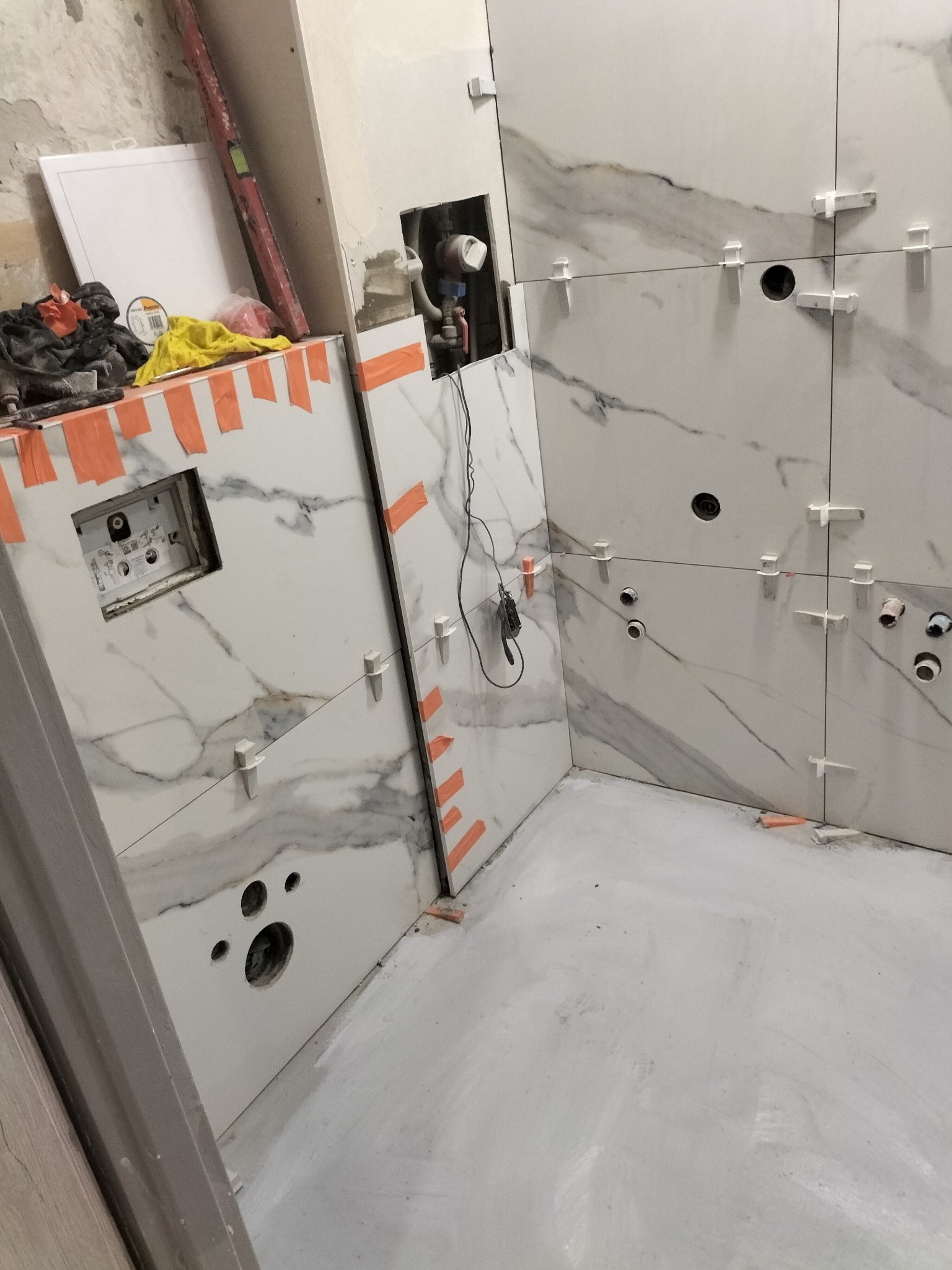 Białe płytki imitujące marmur, ułożone na ścianie łazienki w trakcie prac wykończeniowych; widoczne plastikowe krzyżyki dystansowe, otwory na instalacje i fragment ściany z instalacją podtynkową.