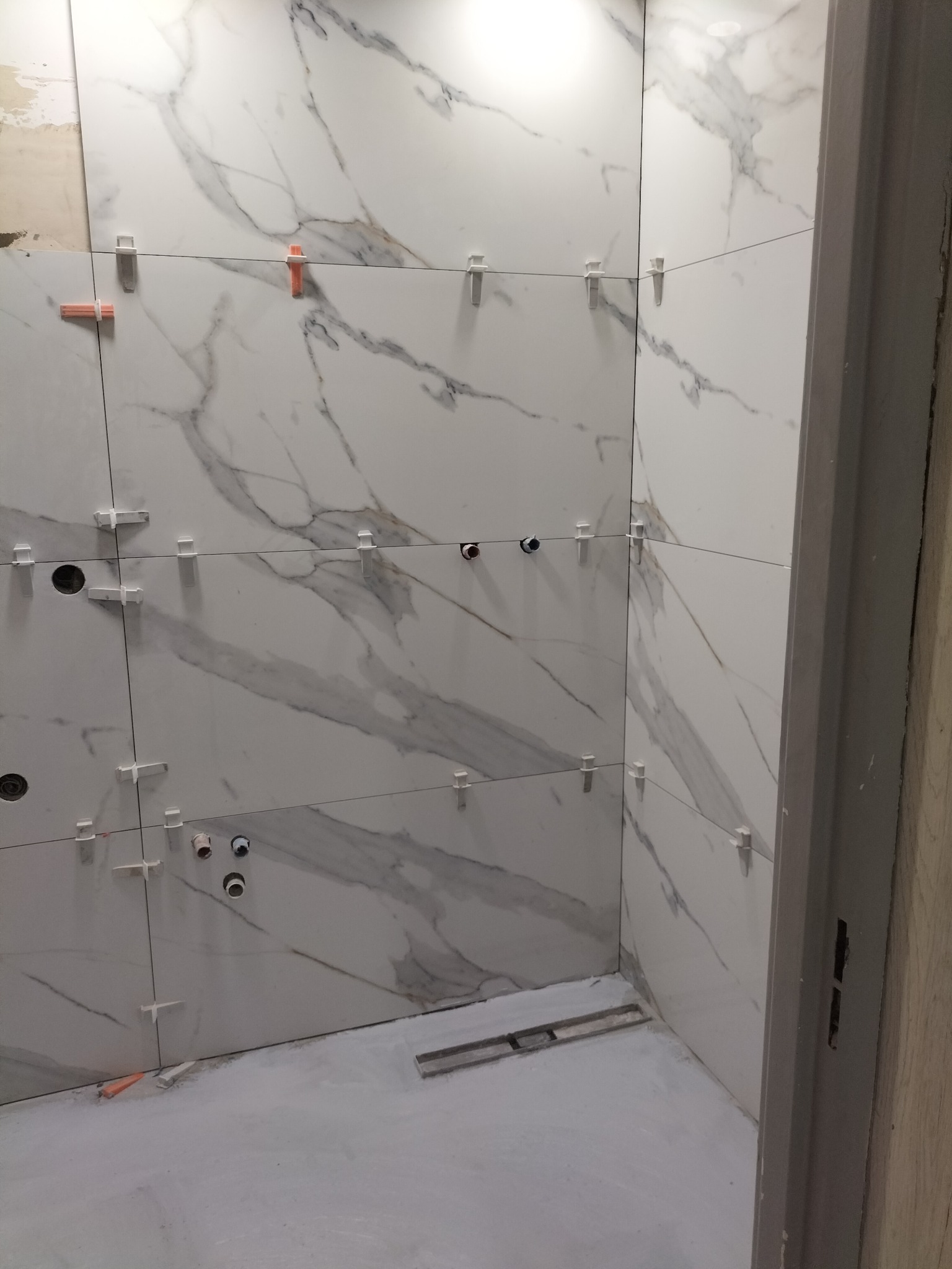 Ściany łazienki w trakcie kładzenia płytek imitujących marmur, z widocznymi plastikowymi krzyżykami dystansowymi i poziomnicą na podłodze.