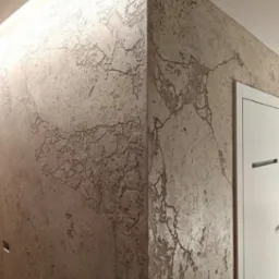 Ściana pokryta dekoracyjną, beżową strukturą przypominającą popękany kamień, oświetlona od góry, z widocznym narożnikiem i fragmentem białych drzwi.