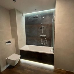 MB FIRMA - Nowoczesna łazienka z podświetlaną wanną i deszczownicą, wykończona płytkami w odcieniach szarości i drewna, z podwieszaną toaletą.