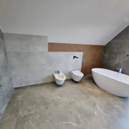 Nowoczesna łazienka na poddaszu z szarymi płytkami imitującymi marmur, białą wanną wolnostojącą, bidetem i podwieszaną toaletą z elementami drewnianej mozaiki na ścianie.