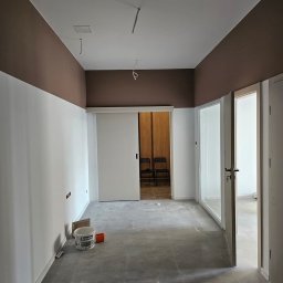 MB FIRMA - Wnętrze po malowaniu: białe ściany z brązowym pasem u góry, szara podłoga, wiadro z farbą Śnieżka obok ściany, przesuwne białe drzwi w głębi