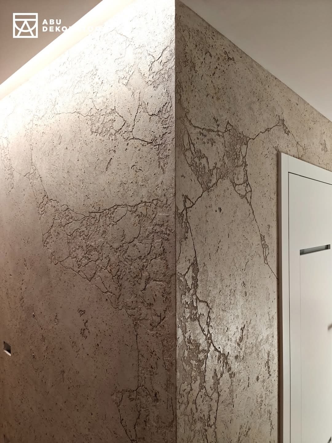 Ściana pokryta dekoracyjną, beżową strukturą przypominającą popękany kamień, oświetlona od góry, z widocznym narożnikiem i fragmentem białych drzwi.