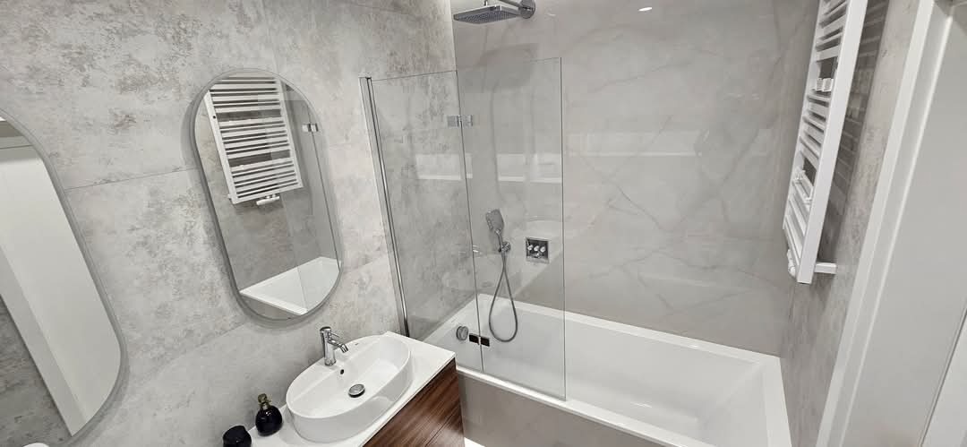 Nowoczesna łazienka z wanną za szklanym panelem, umywalką nablatową na szafce z drewnopodobnym frontem, owalnym lustrem i grzejnikiem drabinkowym na ścianie w odcieniach szarości.