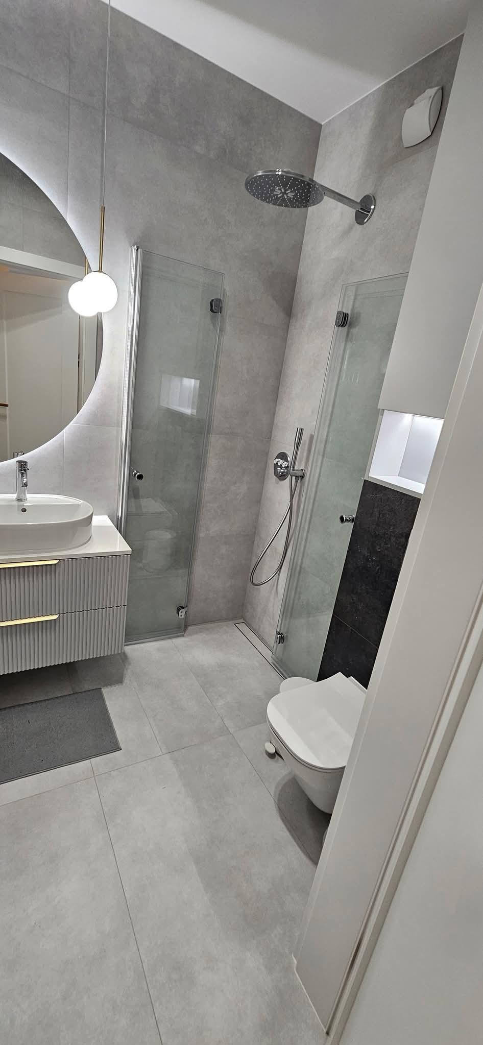 Nowoczesna łazienka w odcieniach szarości z dwoma kabinami prysznicowymi, okrągłym lustrem z podświetleniem LED, umywalką na szafce z ryflowanymi frontami i złotymi uchwytami oraz wiszącą toaletą.