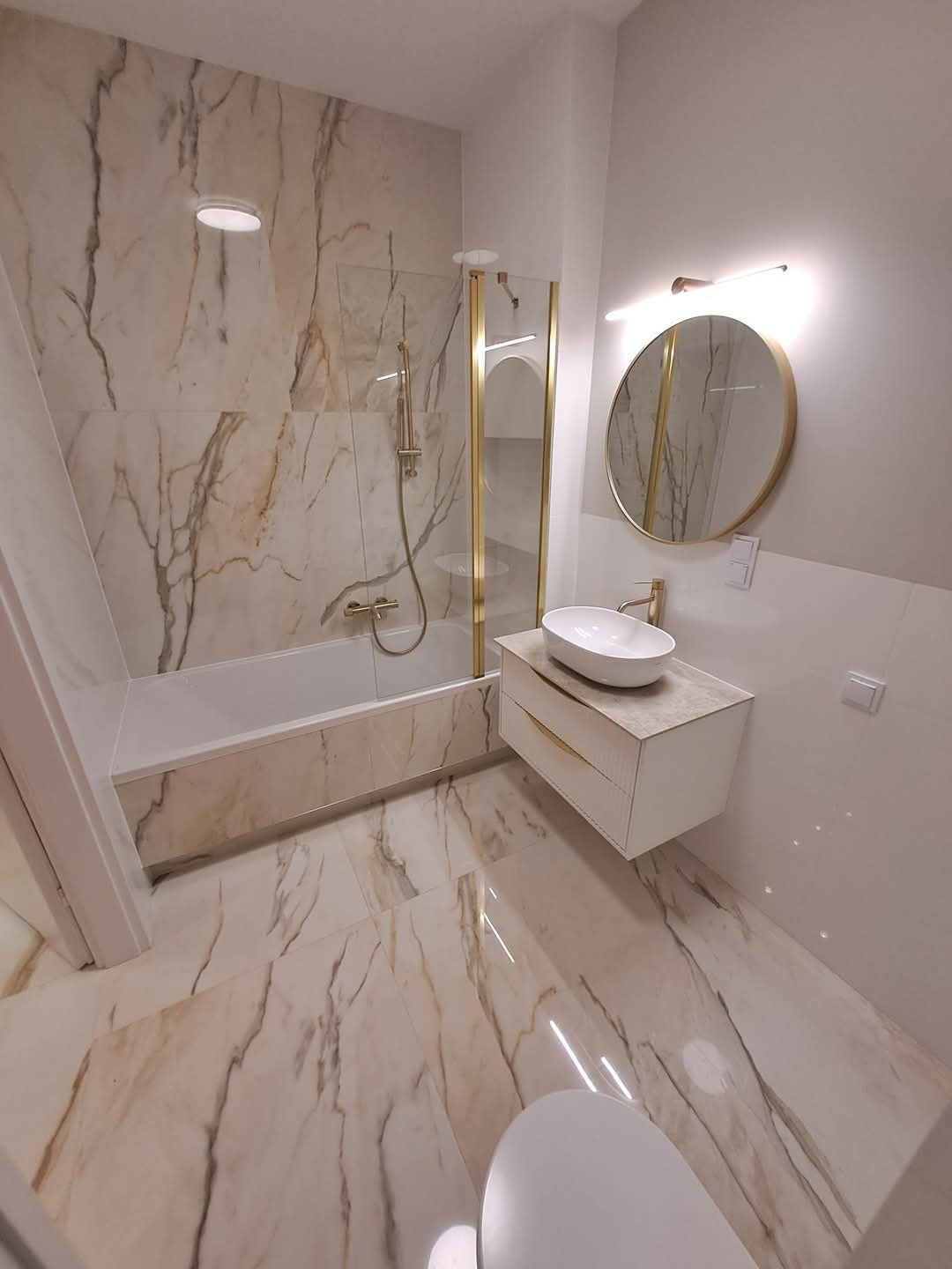 Elegancka łazienka z wanną za szklaną ścianką z elementami w kolorze złota, umywalką nablatową na szafce i okrągłym lustrem z podświetleniem LED.