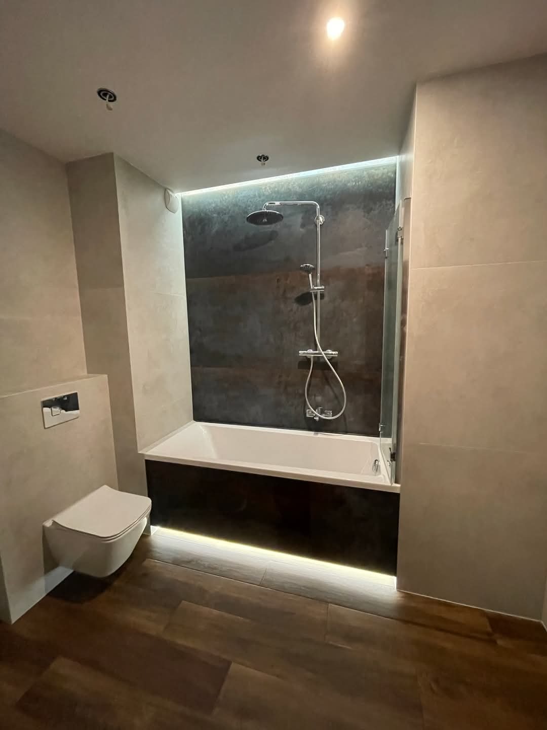 Nowoczesna łazienka z podświetlaną wanną i deszczownicą, wykończona płytkami w odcieniach szarości i drewna, z podwieszaną toaletą.