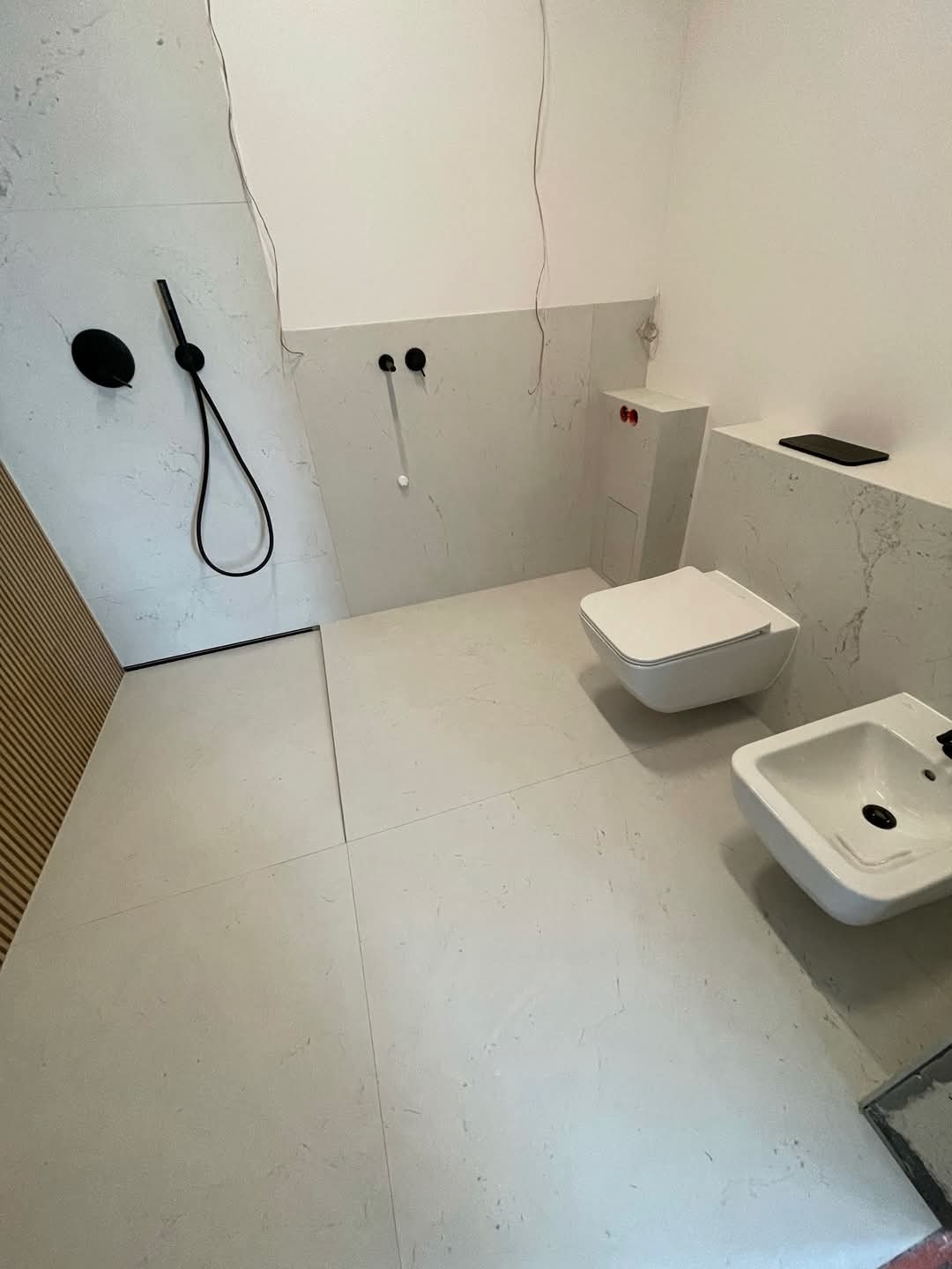 Nowoczesna łazienka w trakcie wykańczania z białymi płytkami imitującymi marmur, wiszącą toaletą i bidetem oraz czarną armaturą prysznicową, widoczne wystające kable i rury