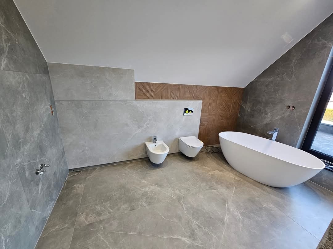 Nowoczesna łazienka na poddaszu z szarymi płytkami imitującymi marmur, białą wanną wolnostojącą, bidetem i podwieszaną toaletą z elementami drewnianej mozaiki na ścianie.