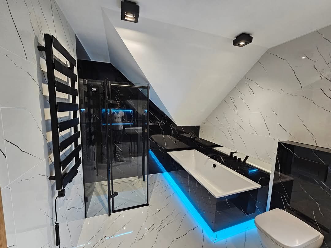 Nowoczesna łazienka na poddaszu z biało-czarnymi marmurkowymi płytkami, podświetlaną na niebiesko wanną, czarną kabiną prysznicową i grzejnikiem drabinkowym.