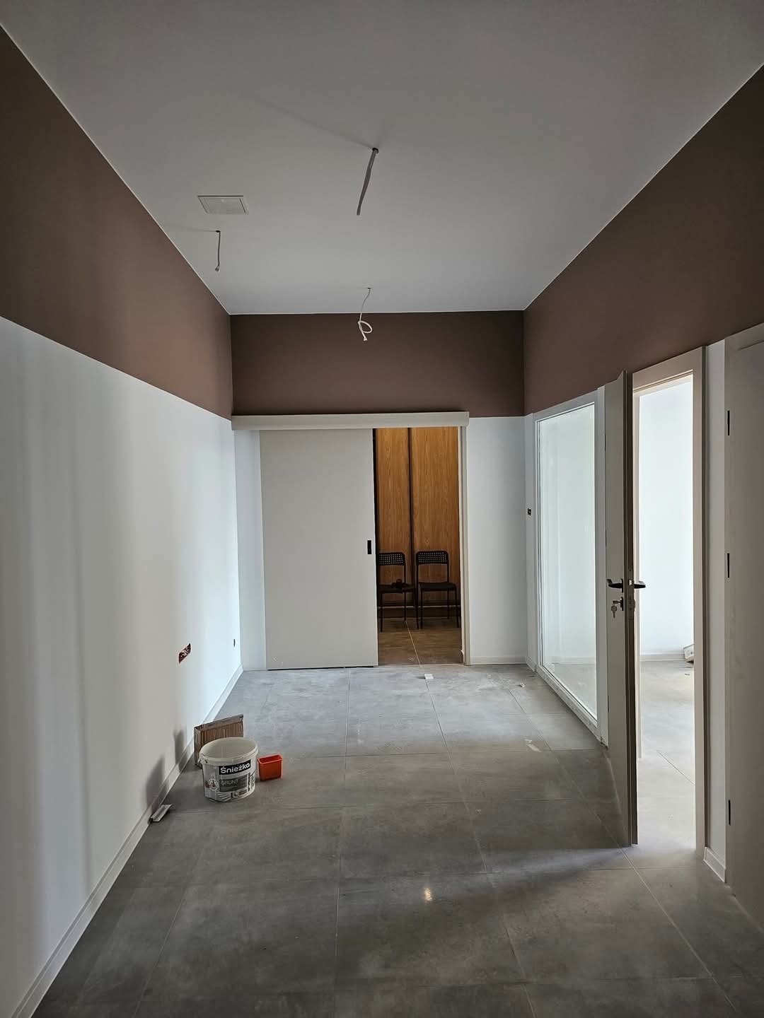 Wnętrze po malowaniu: białe ściany z brązowym pasem u góry, szara podłoga, wiadro z farbą Śnieżka obok ściany, przesuwne białe drzwi w głębi