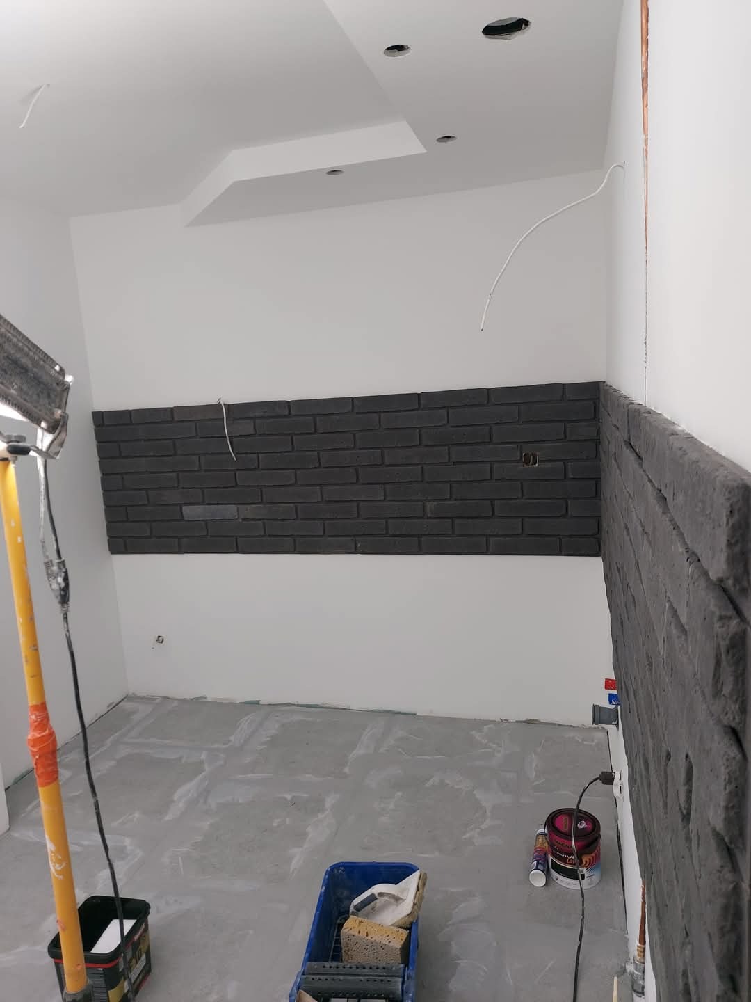 Układanie ciemnoszarej cegły dekoracyjnej na ścianie w trakcie prac wykończeniowych, widoczne narzędzia i materiały budowlane, surowy beton na podłodze, lampa budowlana oświetlająca ścianę.