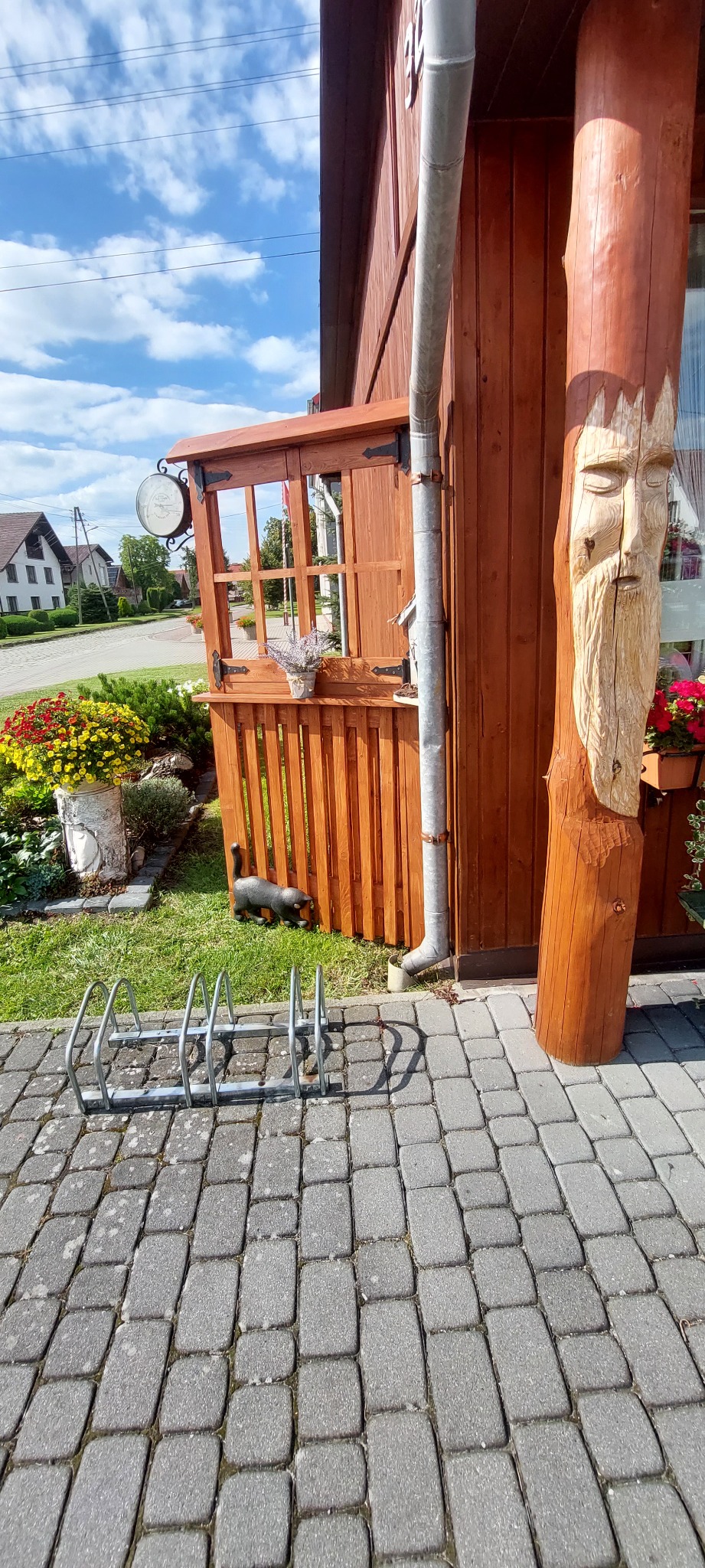 Drewniana dekoracja z rzeźbioną twarzą obok budynku w Osowiec Śląski, widoczne kwiaty w donicach, stojak na rowery i figurka kota na trawniku.