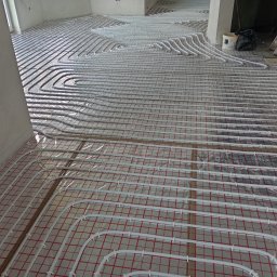 Usługi Hydrauliczne Witold Mac - Rozłożone białe rury ogrzewania podłogowego na czerwonej siatce w surowym wnętrzu, widoczne elementy instalacji i drabina w tle.
