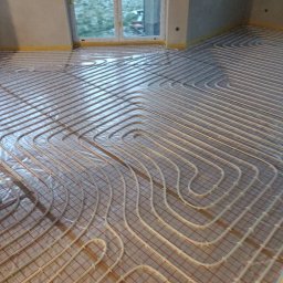 Usługi Hydrauliczne Witold Mac - Instalacja ogrzewania podłogowego w nowym budynku: białe rury ułożone w pętle na izolacji z czerwoną siatką, widok z góry, okno w tle.