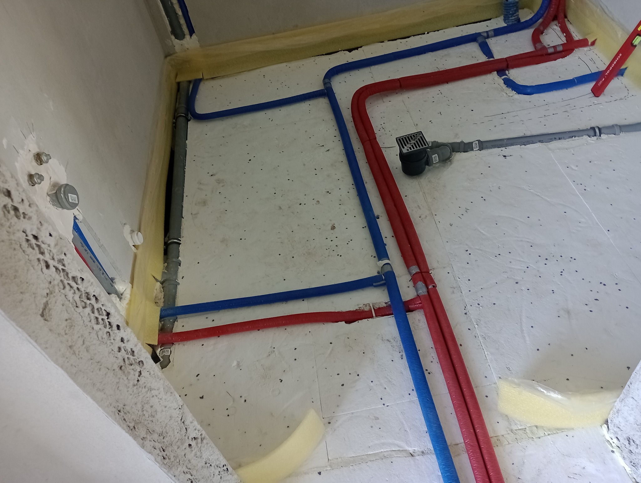 Podłogowe rozprowadzenie instalacji wodnej i grzewczej z rur PEX w kolorze niebieskim i czerwonym, widoczny odpływ liniowy, izolacja termiczna przy ścianach.