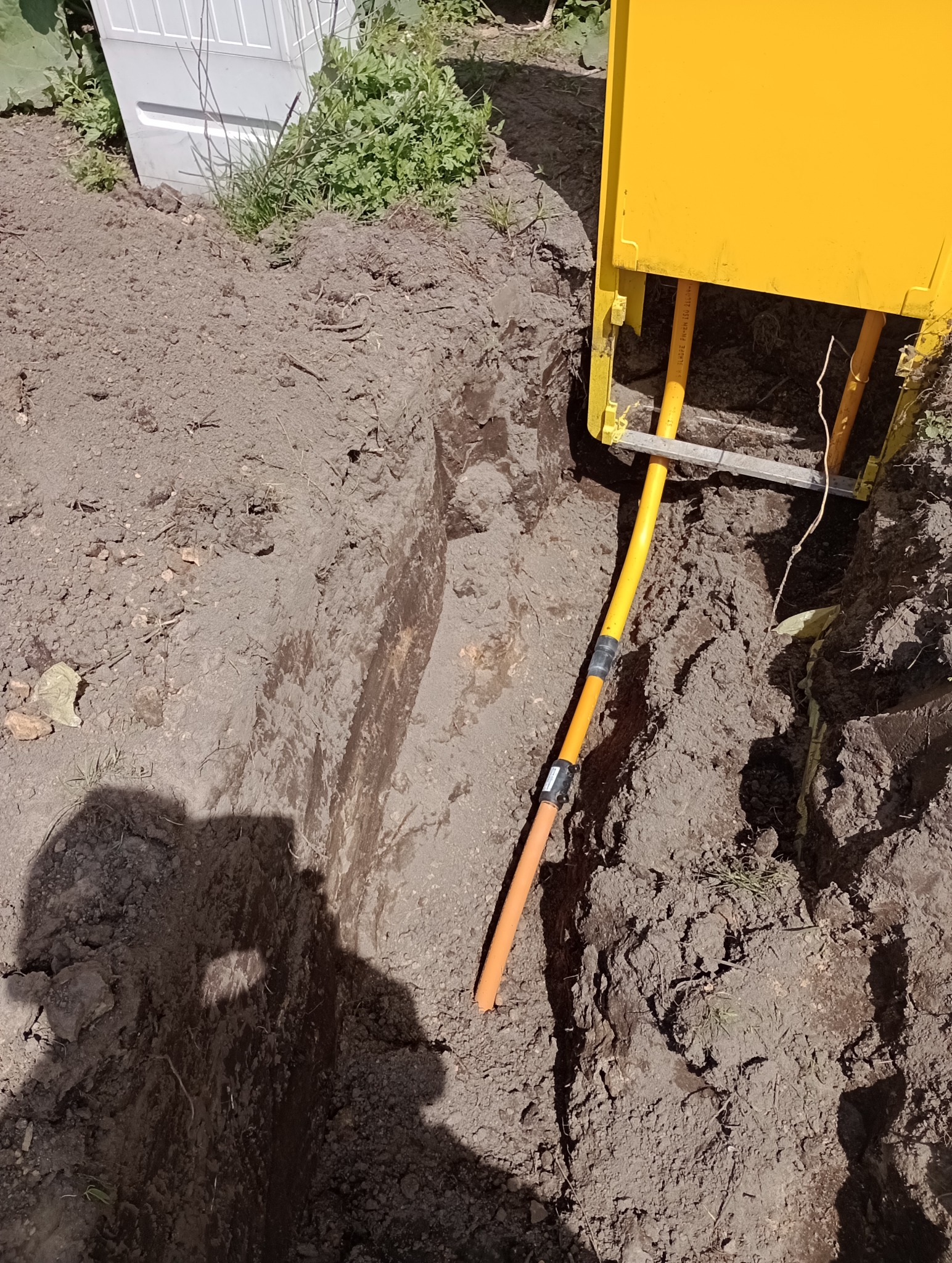 Żółta rura gazowa w wykopie ziemnym, zabezpieczona żółtą skrzynką, widoczny cień osoby.
