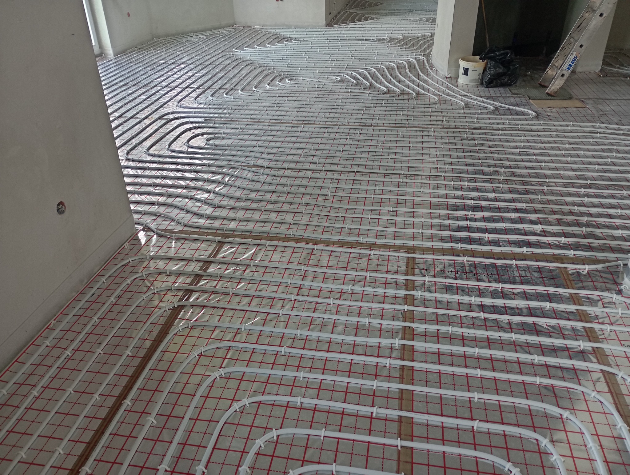 Rozłożone białe rury ogrzewania podłogowego na czerwonej siatce w surowym wnętrzu, widoczne elementy instalacji i drabina w tle.