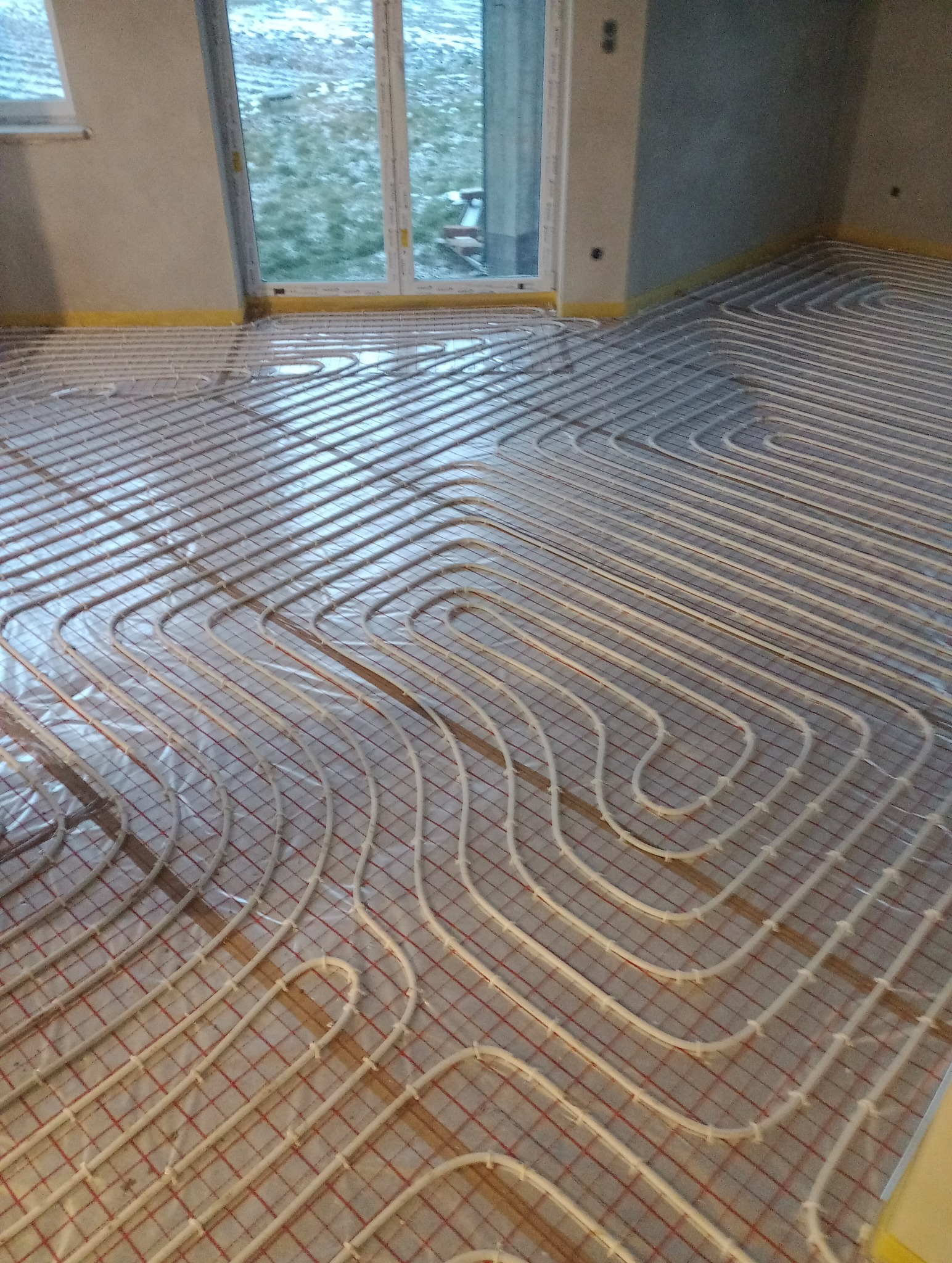 Instalacja ogrzewania podłogowego w nowym budynku: białe rury ułożone w pętle na izolacji z czerwoną siatką, widok z góry, okno w tle.