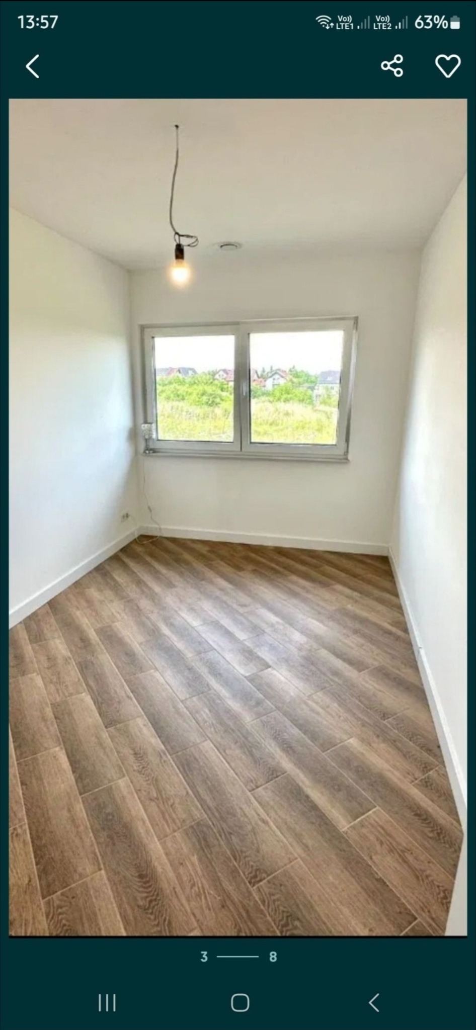 Ukośnie ułożone panele podłogowe w jasnym pokoju z oknem, widok na zieleń za oknem, surowa żarówka na kablu zwisająca z sufitu.