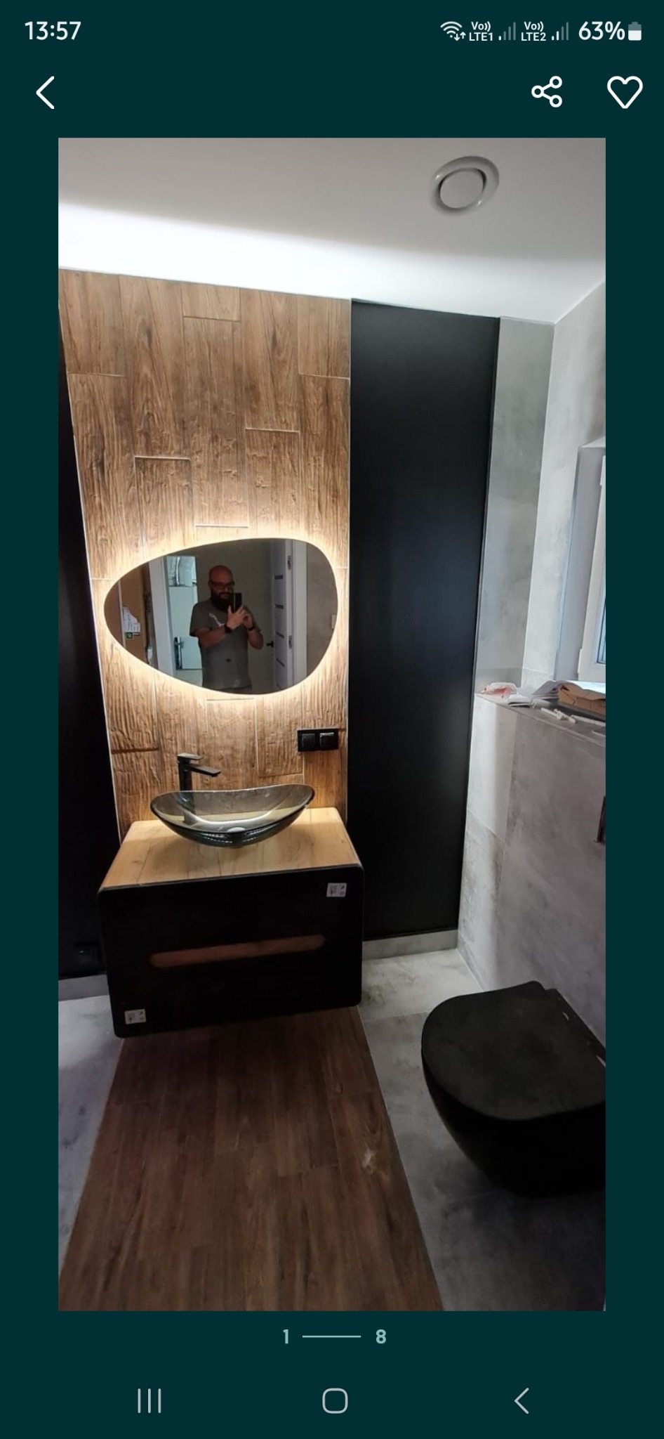 Nowoczesna łazienka z drewnopodobną ścianą, podświetlanym lustrem o nieregularnym kształcie, szklaną umywalką nablatową i czarną armaturą, odbicie fotografa w lustrze.