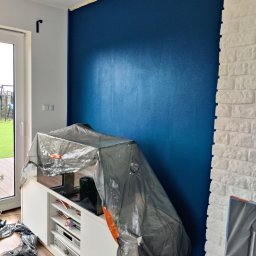 Andrzej Grab - Świeżo pomalowana na granatowo ściana z widocznym odcięciem od sufitu taśmą malarską, zabezpieczony folią mebel, fragment ściany z białej cegły dekoracyjnej.