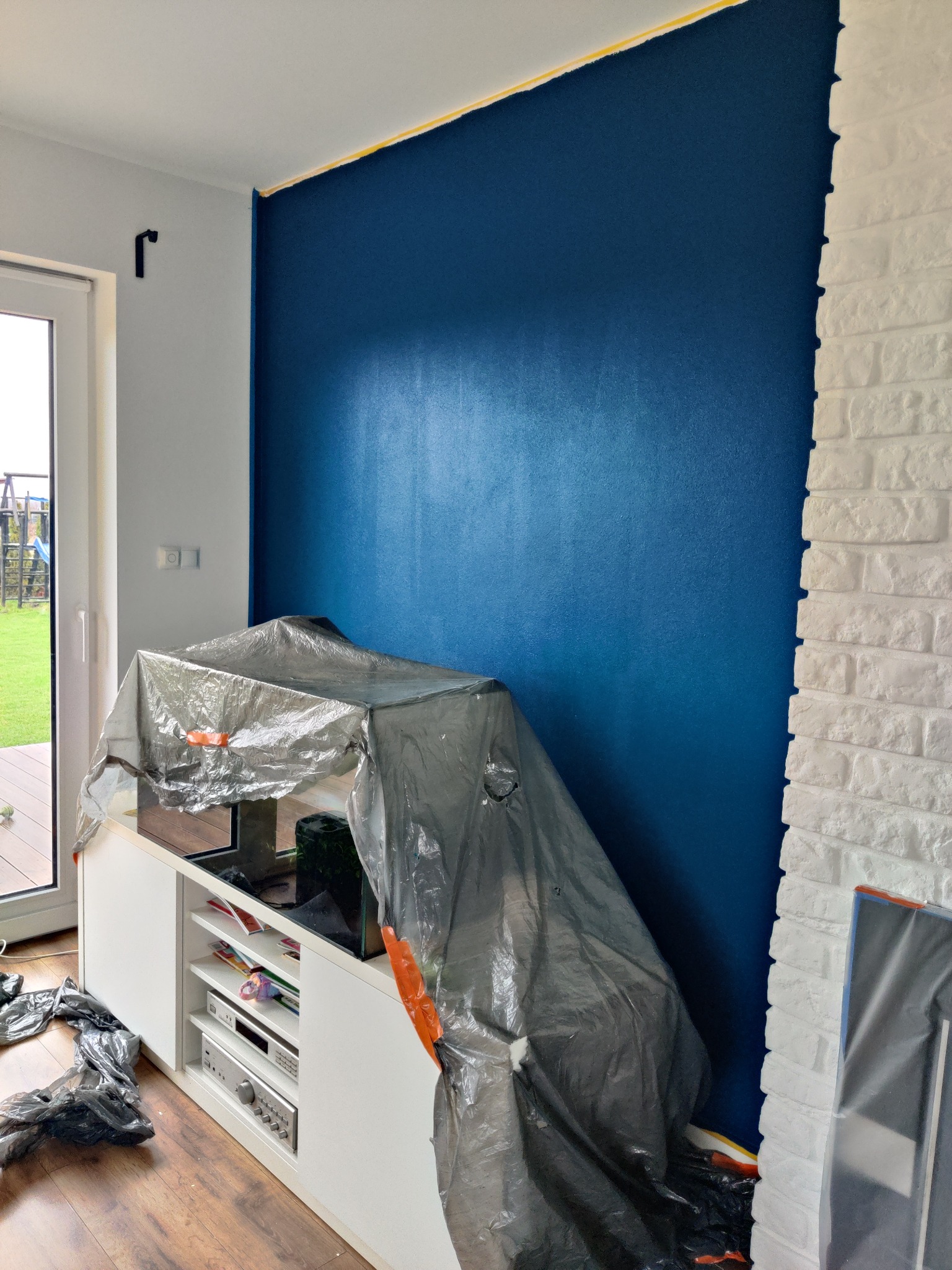 Świeżo pomalowana na granatowo ściana z widocznym odcięciem od sufitu taśmą malarską, zabezpieczony folią mebel, fragment ściany z białej cegły dekoracyjnej.