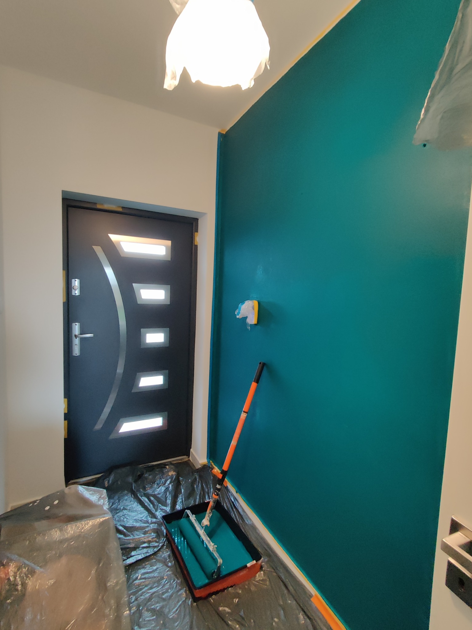 Ściana w odcieniu turkusu, świeżo pomalowana, z wałkiem i kuwetą na folii ochronnej, widoczna nowoczesna czarna stolarka drzwiowa z metalową aplikacją.
