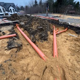 ALL INVESTMENTS SPÓŁKA KOMANDYTOWA - Instalacja kanalizacyjna z pomarańczowych rur PVC w piasku, częściowo zakopana, na budowie domu jednorodzinnego. W tle fundamenty i osoba w roboczym ubraniu.