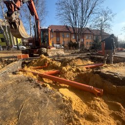 ALL INVESTMENTS SPÓŁKA KOMANDYTOWA - Pomarańczowe rury w wykopie, koparka w tle. Prace ziemne przy instalacji wodno-kanalizacyjnej. Widoczny pracownik i budynki w oddali.
