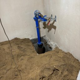 ALL INVESTMENTS SPÓŁKA KOMANDYTOWA - Niebieskie przyłącze wodne z licznikiem, zamontowane w piwnicy, z widocznym wykopem i piaskiem. Instalacja zabezpieczona wspornikami przy ścianie.