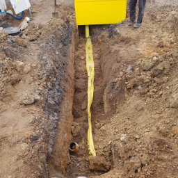 ALL INVESTMENTS SPÓŁKA KOMANDYTOWA - Wykop z żółtą rurą osłonową i taśmą ostrzegawczą w ziemi, obok żółta skrzynka i narzędzia. Widoczny fragment pracownika w kombinezonie.