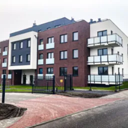 Nowoczesny budynek wielorodzinny z balkonami i ogrodzeniem, widok z zewnątrz, kostka brukowa na podjeździe, elementy ceglane i białe wykończenie.