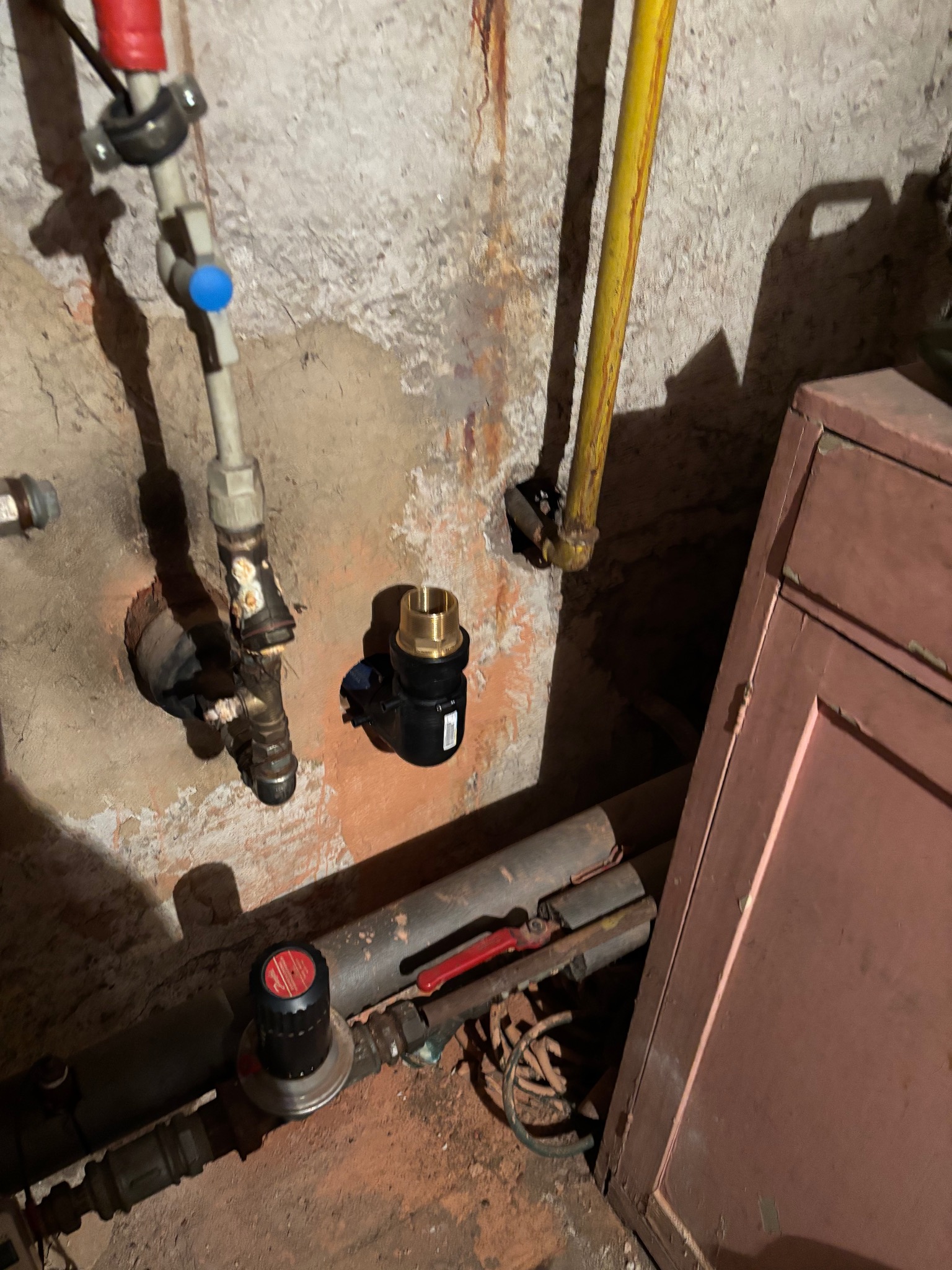 Fragment instalacji hydraulicznej przy starej ścianie z widocznymi rurami, zaworami i nowym przyłączem. Nierówności ściany i oświetlenie podkreślają surowy charakter.