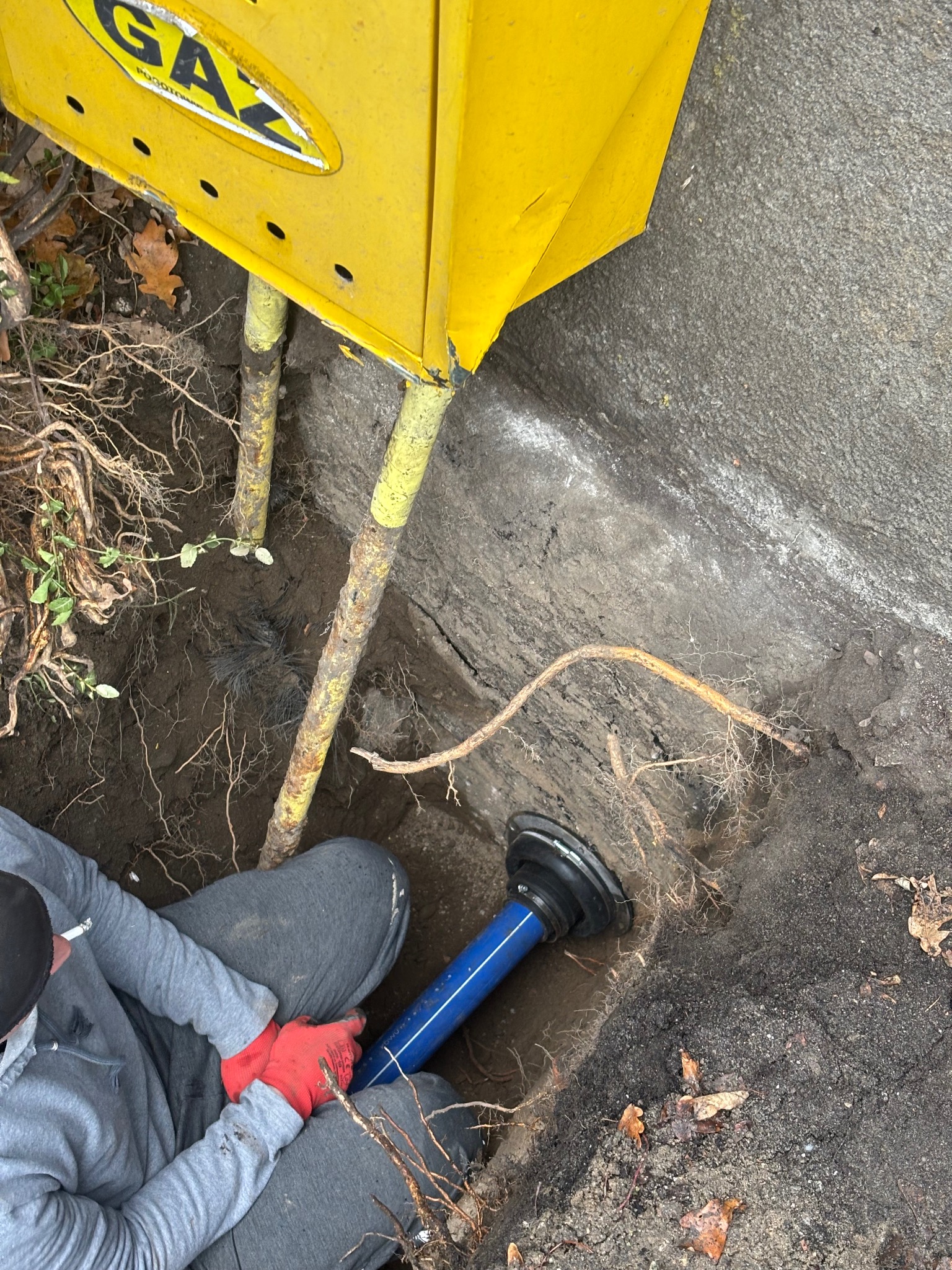Pracownik w wykopie łączy niebieską rurę z żółtą skrzynką gazową. Widoczne korzenie, ziemia i fragment betonowej ściany. Czerwone rękawice robocze.