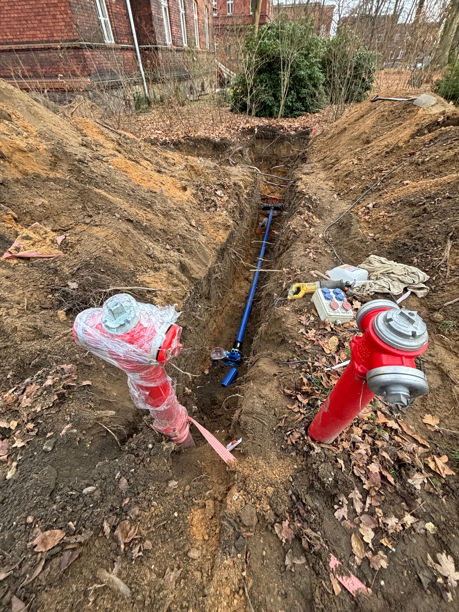 Wykonanie przyłącza wodociągowego: wykop z rurą w ziemi, dwa czerwone hydranty po bokach, jeden z nich owinięty folią ochronną. Widoczne narzędzia i kable.