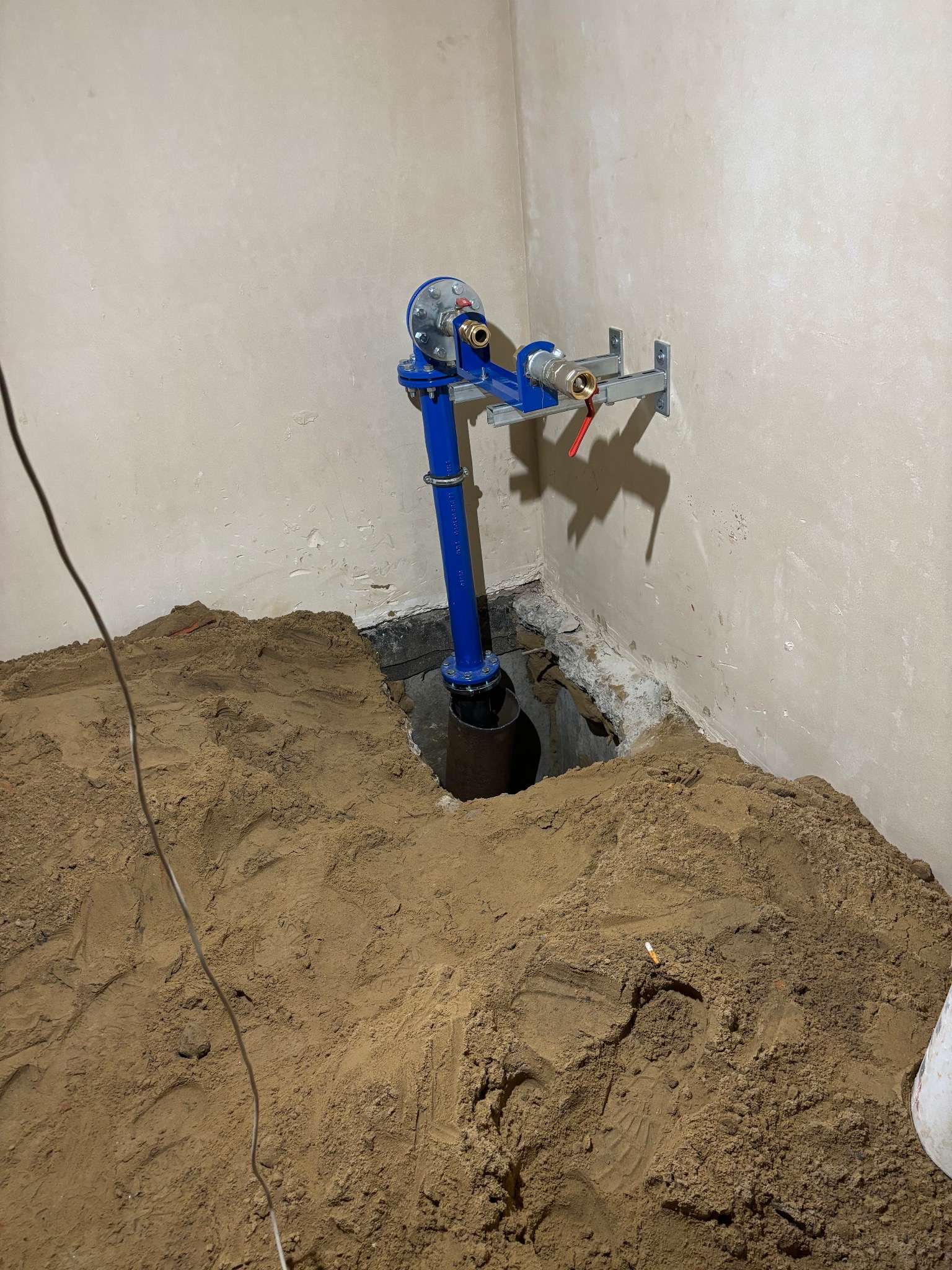 Niebieskie przyłącze wodne z licznikiem, zamontowane w piwnicy, z widocznym wykopem i piaskiem. Instalacja zabezpieczona wspornikami przy ścianie.