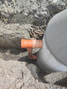 Pomarańczowe rury kanalizacyjne podłączone do betonowej studni w wykopie ziemnym. Widoczne połączenia i fragment studni. Drobne kamienie i gruz w tle.