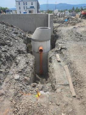 Betonowy zbiornik z rurą kanalizacyjną w wykopie, widok z góry, w tle budynek i góry. Element instalacji sanitarnej w trakcie budowy.