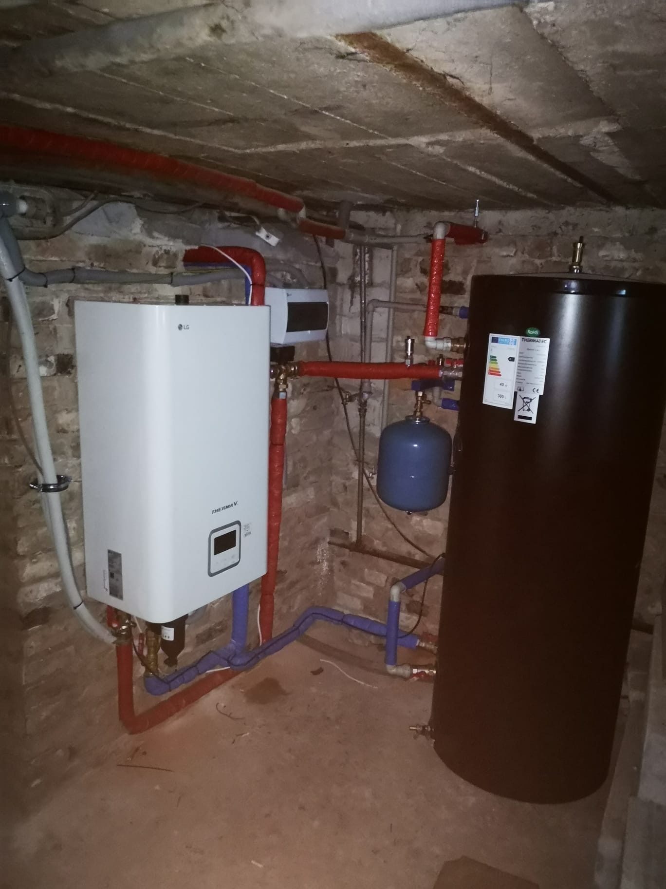 Instalacja CO w piwnicy: biały piec LG Therma V, czarny zbiornik, niebieski zbiornik wyrównawczy, czerwone i niebieskie rury. Surowy beton i cegła w tle.