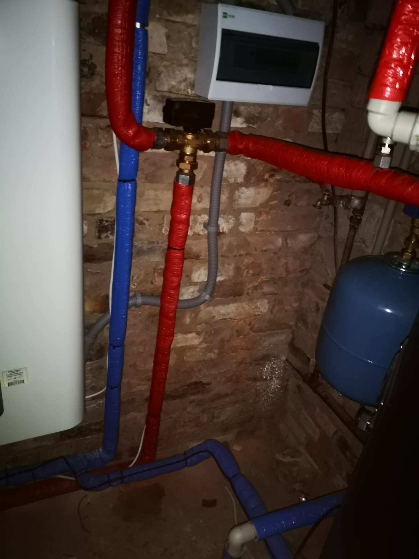 Instalacja grzewcza z rurami w izolacji (czerwona i niebieska) na tle ceglanej ściany. Widoczny biały bojler i szare rury.