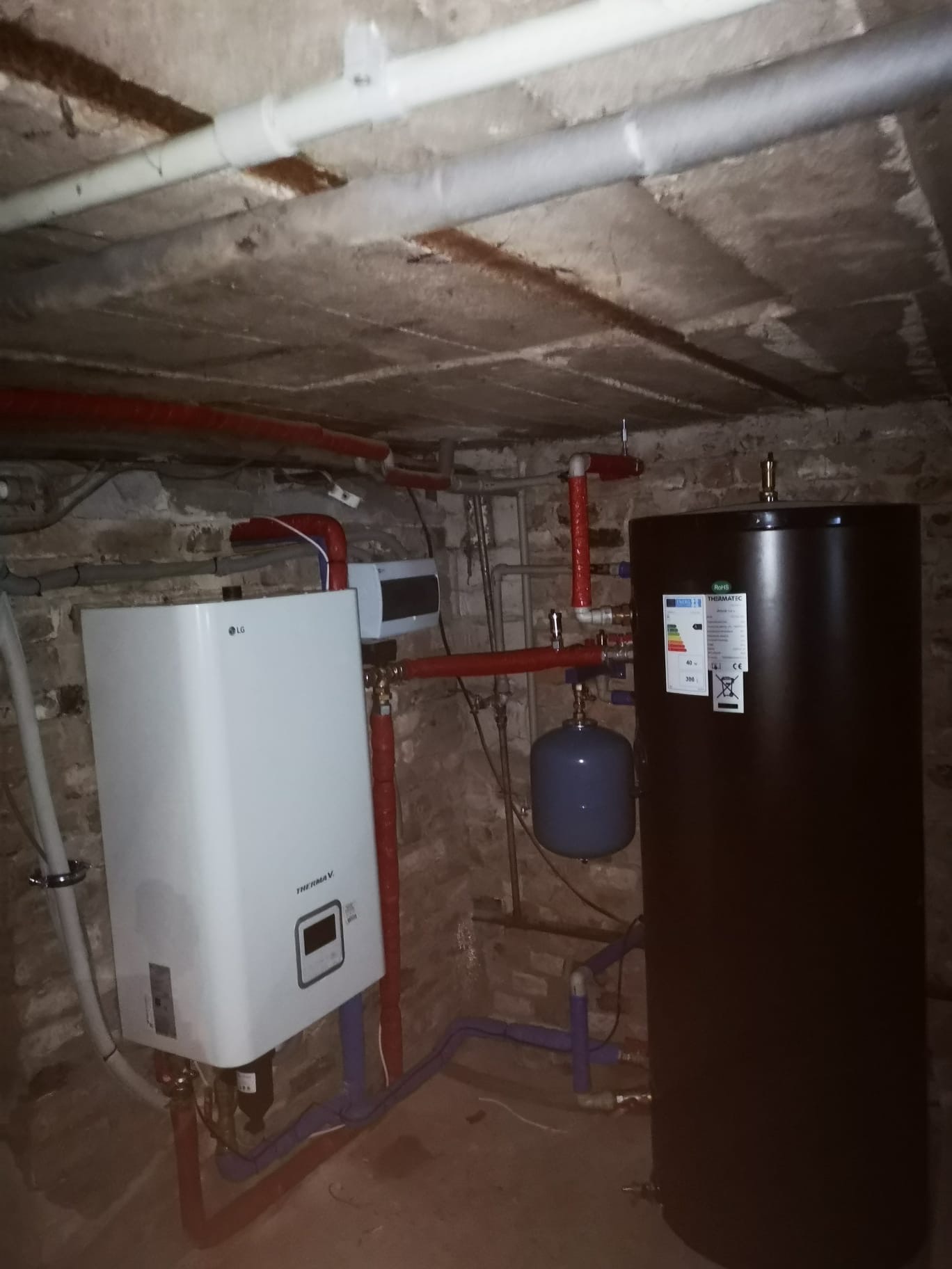 Kotłownia z białym piecem LG Therma V i czarnym zasobnikiem Thermatec. Instalacja z rur w kolorze czerwonym i fioletowym. Surowy betonowy strop.