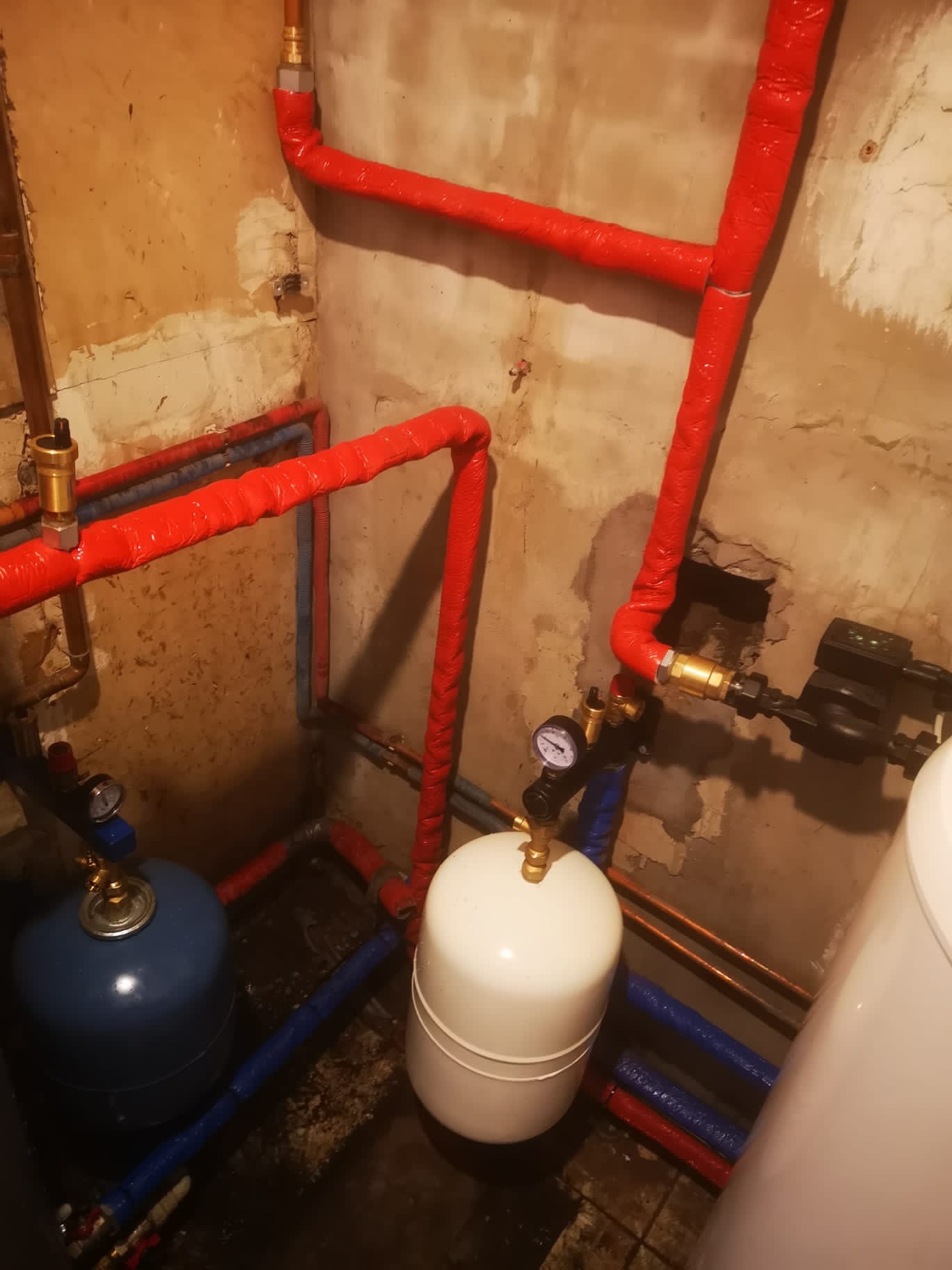 Zabezpieczone rury grzewcze w izolacji czerwonej, zbiorniki wyrównawcze: biały i niebieski, manometr, instalacja wodna i grzewcza w kotłowni.