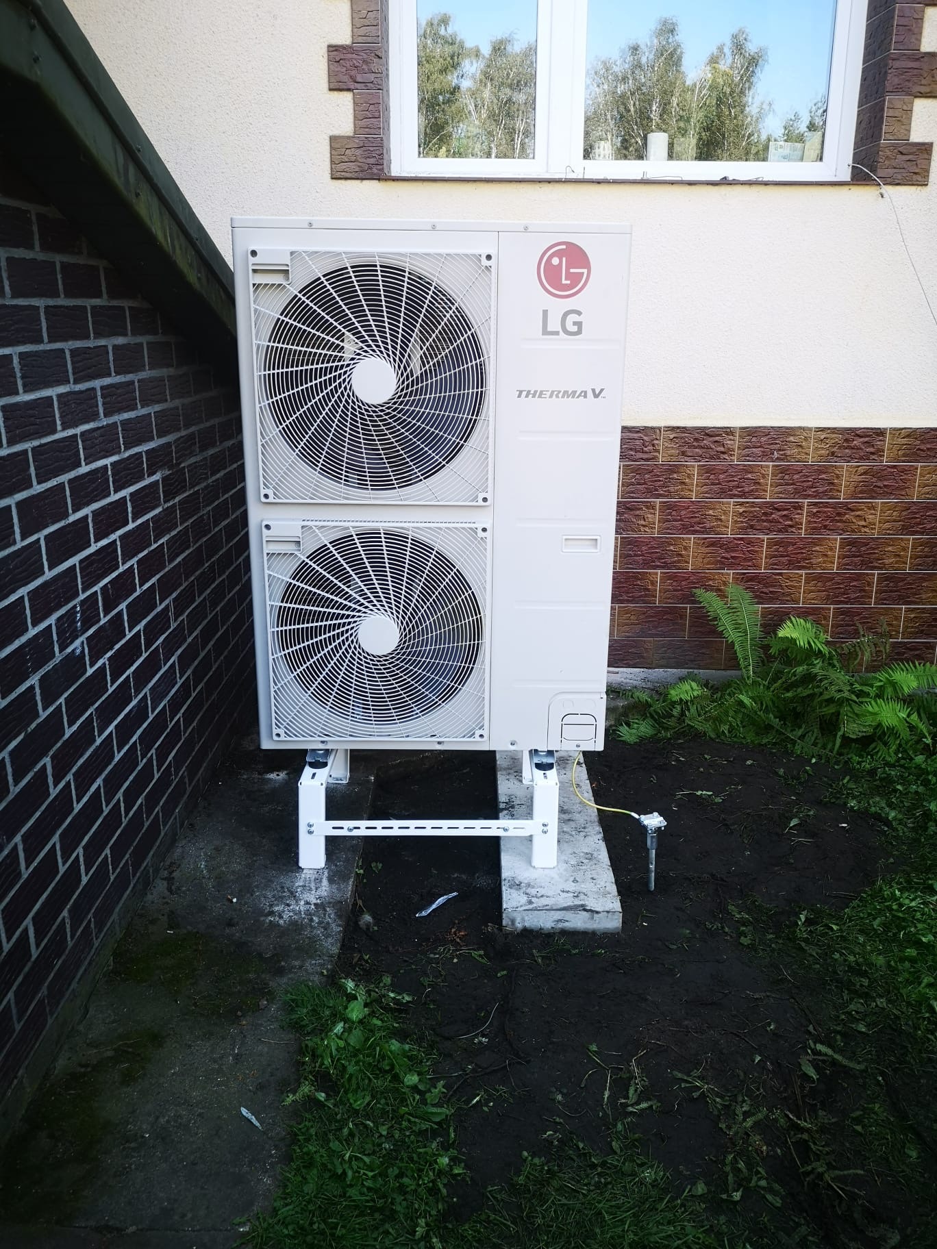 Biała pompa ciepła LG Therma V, zamontowana na betonowym fundamencie, z widocznymi dwoma wentylatorami i instalacją uziemiającą, na tle ściany budynku.