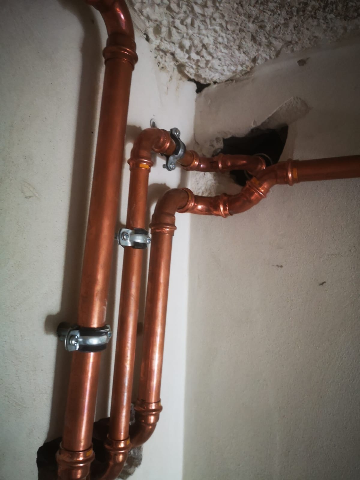 Miedziane rury instalacji, mocowane do ściany klamrami, wewnątrz pomieszczenia. Widoczne połączenia lutowane i fragment sufitu z ubytkiem tynku.