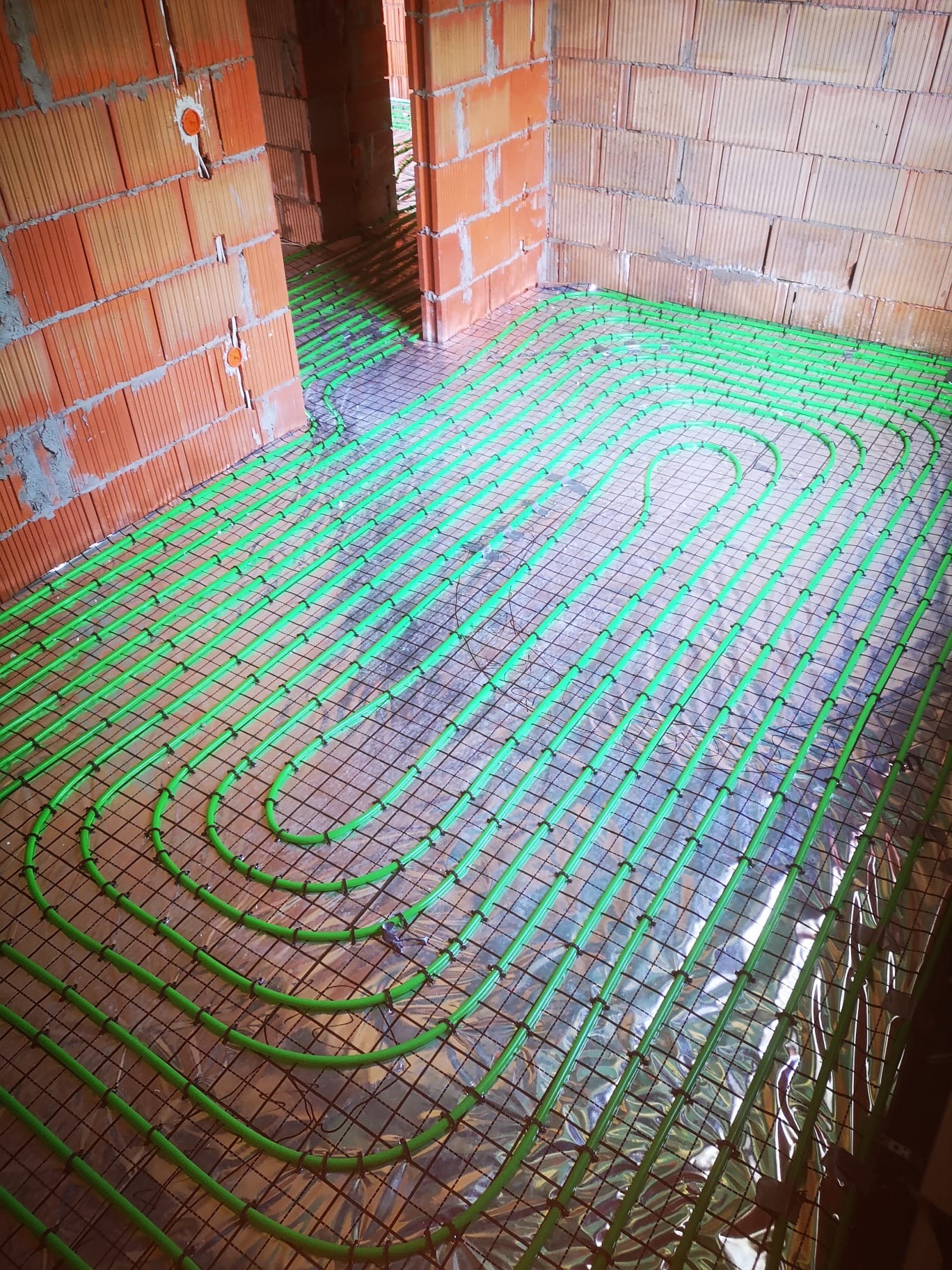 Zielone rury ogrzewania podłogowego, zamontowane na folii termoizolacyjnej i siatce, w surowym wnętrzu domu w Chorzowie. Instalacja przed wylaniem posadzki.