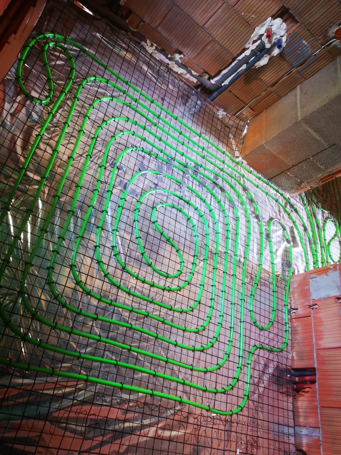 Ściana z cegły z zamontowanym systemem ogrzewania podłogowego w postaci zielonych rurek ułożonych spiralnie na metalowej siatce. Widoczne rury instalacji pod sufitem.